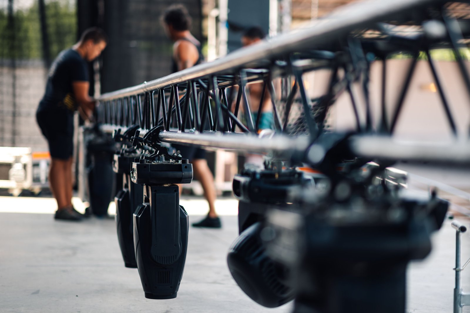 Several stage lights hanging from an aluminum truss system are being prepared for a live music concert or other event by stage technicians